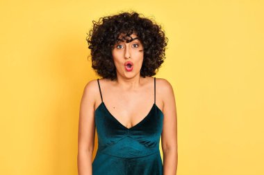 Young arab woman with curly hair wearing elegant dress over isolated yellow background afraid and shocked with surprise expression, fear and excited face.