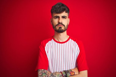 İzole edilmiş kırmızı arka plan üzerinde duran çizgili tişörtlü genç bir adam şüpheci ve gergin, yüzünde çapraz kollu tasvip etmeyen bir ifade. Negatif kişi.