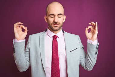 Takım elbiseli ve kravatlı genç bir iş adamı mor izole edilmiş arka planda rahatlar ve parmaklarıyla meditasyon yaparken gözleri kapalı gülümser. Yoga konsepti.