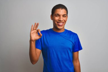 Mavi tişört giyen yakışıklı Arap adam izole edilmiş beyaz arka planda durup gülümsüyor ve el ve parmaklarıyla tamam işareti yapıyor. Başarılı ifade.