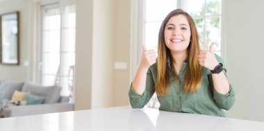 Evdeki genç ve güzel bir kadın. Başparmağıyla gülümseyen ve mutlu bir şekilde olumlu jest yapıyor. Kameraya neşeli bir ifadeyle bakmak, kazanan bir jest..