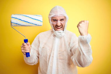 Boyacı kıyafeti giyen genç adam, izole edilmiş sarı arka planda boya rulosu tutuyor öfkeyle bağırıyor, deliriyor ve el kaldırarak bağırıyor. Öfke kavramı.