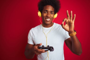 Afro oyuncu video oyunu oynuyor. İzole edilmiş kırmızı arkaplan üzerinde joystick kulaklık kullanıyor. Tamam işareti yapıyor.