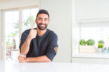 Handsome hispanic man wearing casual sweater at home doing happy thumbs up gesture with hand. Approving expression looking at the camera showing success.