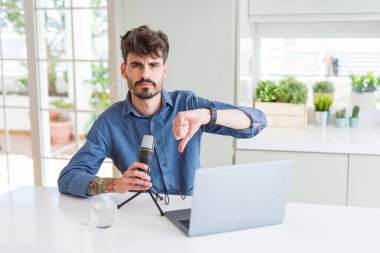 Genç adam mikrofon ve dizüstü bilgisayarla podcast kaydediyor. kızgın yüzlü, negatif işaretli başparmaklar aşağı, reddedilme konsepti