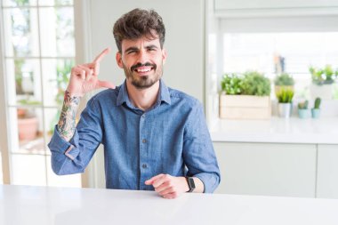 Genç adam beyaz bir masada oturmuş gülümsüyor ve kendine güvenen hareketlerle parmaklarıyla işaret yapıyor ve kameraya bakıyor. Ölçü kavramı.