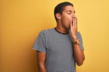 İzole edilmiş sarı arka plan üzerinde denizci çizgili tişört giyen genç, yakışıklı Arap adam esnemekten sıkılmış, ağzı eliyle kapatmaktan sıkılmış. Huzursuz ve uykulu..