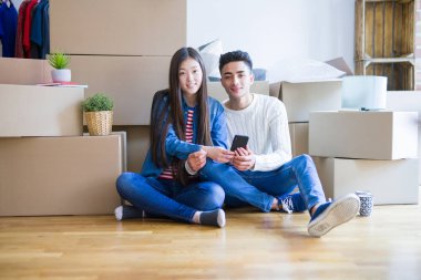 Genç Asya çift oturan üzerinde bu yeni daire civarı karton kutular, kullanarak Smartphone ve gülümsüyor yeni ev