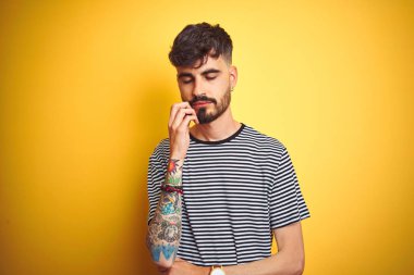 Çizgili t-shirt giyen dövme giyen genç adam izole sarı arka plan üzerinde duran yorgun görünümlü ve çapraz kolları ile depresyon sorunları ile sıkılmış düşünme.