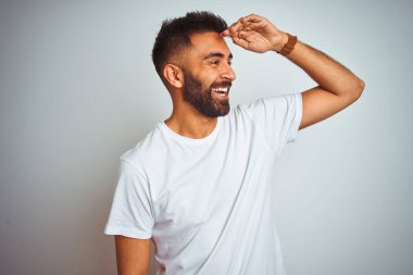 Young indian man wearing t-shirt standing over isolated white background very happy and smiling looking far away with hand over head. Searching concept.