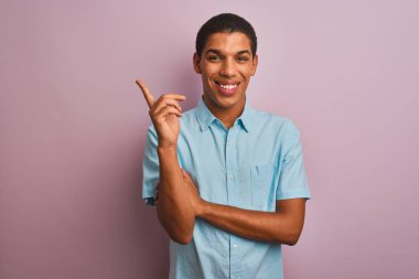 Mavi gömlekli genç yakışıklı Arap adam pembe arka planda duruyor yüzünde kocaman bir gülümseme, eli ve parmağı kameraya bakıyor..