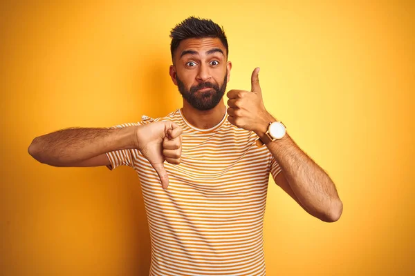 孤立した黄色の背景の上に立つTシャツを着た若いインド人男性親指を上下に 意見の相違や合意表現を行います 狂気の葛藤 — ストック写真