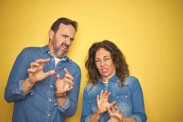 Beautiful middle age couple together standing over isolated yellow background disgusted expression, displeased and fearful doing disgust face because aversion reaction. With hands raised. Annoying concept.
