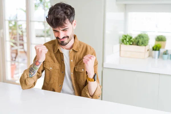 Young man wearing casual jacket sitting on white table very happy and excited doing winner gesture with arms raised, smiling and screaming for success. Celebration concept.