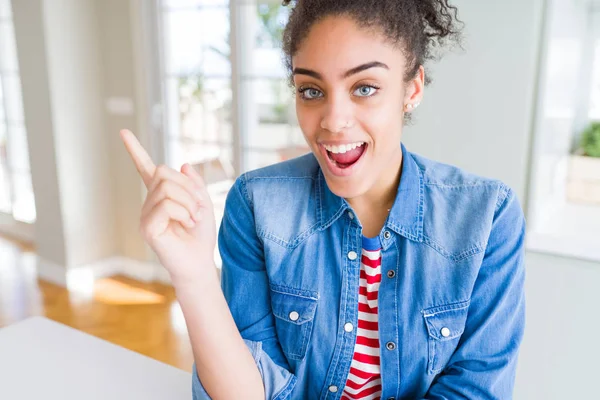 Beautiful young african american woman with afro hair wearing casual denim jacket with a big smile on face, pointing with hand and finger to the side looking at the camera.