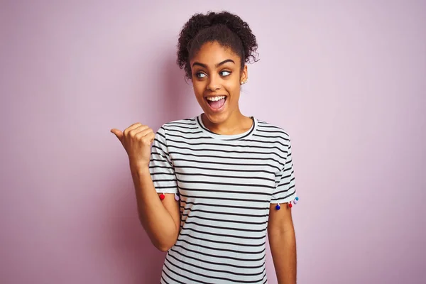 African american woman wearing navy striped t-shirt standing over isolated pink background smiling with happy face looking and pointing to the side with thumb up.