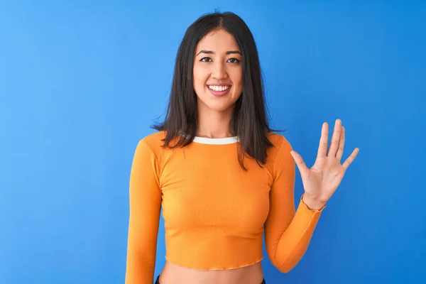 年轻美丽的中国女人穿着橙色的T恤 站在孤零零的蓝色背景上 带着五号手指指尖 满面笑容 自信而快乐 — 图库照片