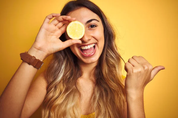 年轻而美丽的女人 用一只柠檬挡住黄色的孤立的背景 用拇指指向旁边 笑容满面 — 图库照片