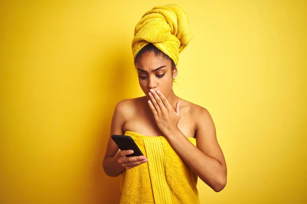 Afro woman wearing towel after shower using smatrphone over isolated yellow background cover mouth with hand shocked with shame for mistake, expression of fear, scared in silence, secret concept