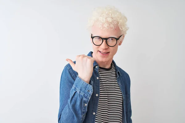 Young albino blond man wearing denim shirt and glasses over isolated white background smiling with happy face looking and pointing to the side with thumb up.