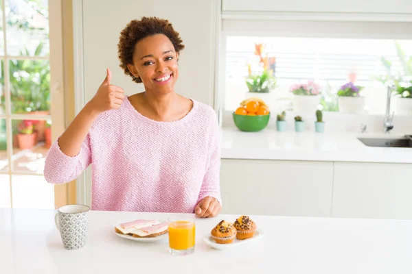Young african american woman eating breaksfast in the morning at home doing happy thumbs up gesture with hand. Approving expression looking at the camera with showing success.