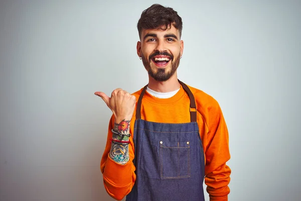 Young shopkeeper man with tattoo wearing apron standing over isolated white background smiling with happy face looking and pointing to the side with thumb up.