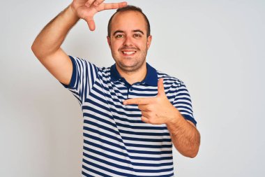 Sıradan çizgili polo giyen genç adam izole edilmiş beyaz arka planda durup gülümsüyor ve mutlu yüzlü elleri ve parmakları olan bir çerçeve yapıyor. Yaratıcılık ve fotoğrafçılık kavramı.