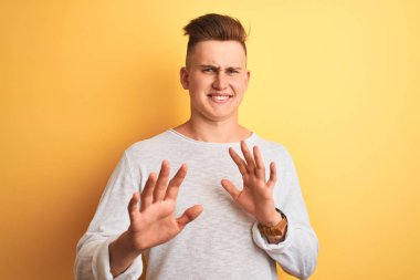 İzole edilmiş sarı arka plan üzerinde duran beyaz tişörtlü genç yakışıklı adam tiksindirici ve tiksindirici bir yüz ifadesi takınıyor çünkü tiksinti tepkisi veriyor. Eller havada. Can sıkıcı bir kavram..