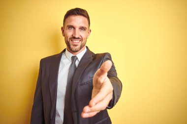 Sarı arka plan üzerinde genç ve yakışıklı bir iş adamı gülümseyerek selamlama ve hoş geldin demek için tokalaşıyor. Başarılı bir iş..