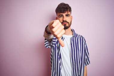 İzole edilmiş pembe arka plan üzerinde duran çizgili tişörtlü genç adam mutsuz ve kızgın görünüyor. Reddedilmiş ve olumsuz jestler yapıyor. Kötü ifade.