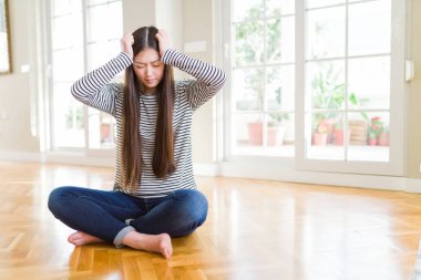 Beautiful Asian woman sitting barefoot on the floor at home suffering from headache desperate and stressed because pain and migraine. Hands on head.
