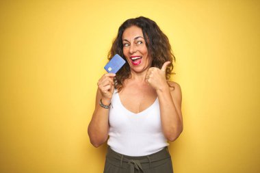 Orta yaşlı yaşlı bir kadın kredi kartını sarı arka planda tutuyor ve gülümseyen bir gülümsemeyle baş parmağıyla işaret ediyor.