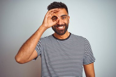 Young indian man wearing black striped t-shirt standing over isolated white background doing ok gesture with hand smiling, eye looking through fingers with happy face.