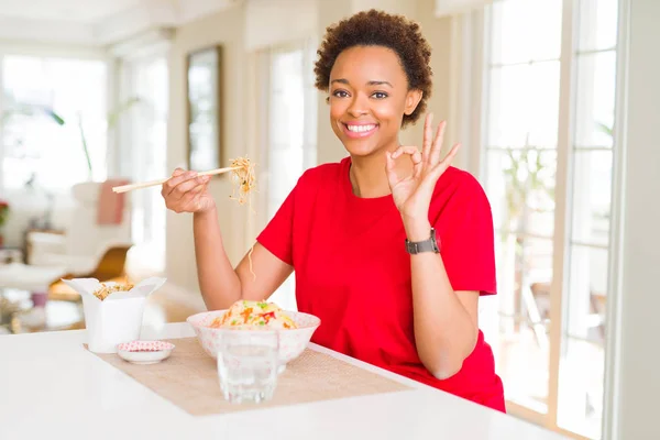 Young african american woman with afro hair eating asian food at home doing ok sign with fingers, excellent symbol