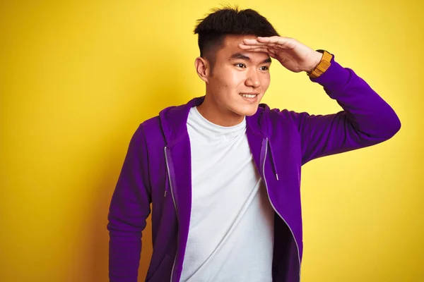 孤立した黄色の背景の上に立って紫色のスウェットシャツを着て若いアジア系中国人男性は非常に幸せと笑みを浮かべて遠くに手を頭の上に見て 検索の概念 — ストック写真