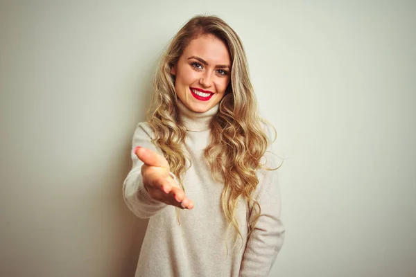 Beautiful woman wearing winter turtleneck sweater over isolated white background smiling friendly offering handshake as greeting and welcoming. Successful business.