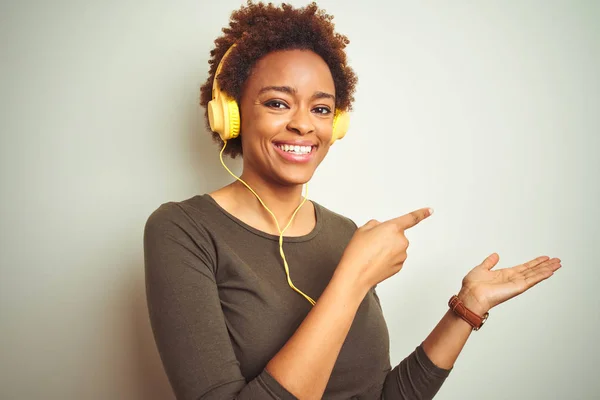 African american woman wearing headphones listening to music over isolated background amazed and smiling to the camera while presenting with hand and pointing with finger.