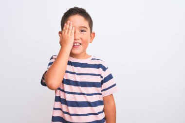 Güzel çocuk, günlük çizgili tişört giyiyor. İzole edilmiş beyaz arka plan üzerinde duruyor. Bir gözünü eliyle kapatıyor..