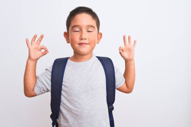 Güzel öğrenci çocuk sırt çantası takıp izole edilmiş beyaz arka plan üzerinde duruyor rahatlıyor ve parmaklarıyla meditasyon hareketi yaparken gözleri kapalı gülümsüyor. Yoga konsepti.