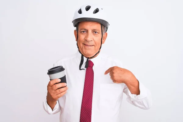 Senior businessman wearing bike helmet drinking coffee over isolated white background with surprise face pointing finger to himself