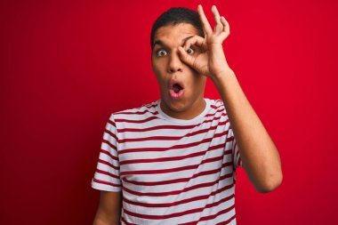 İzole edilmiş kırmızı arka plan üzerinde çizgili tişört giyen genç, yakışıklı Arap adam şaşırmış yüz ifadesiyle şaşırmış, parmaklarından bakan bir hareket yapıyor. İnançsız ifade.