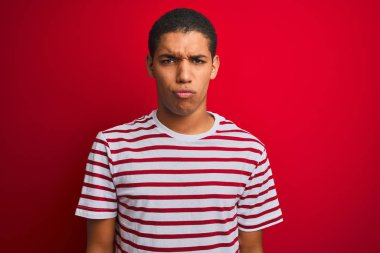 İzole edilmiş kırmızı arka plan üzerinde çizgili tişört giyen genç yakışıklı Arap adam şüpheci ve gergin, sorun yüzünden surat asıyor. Negatif kişi.