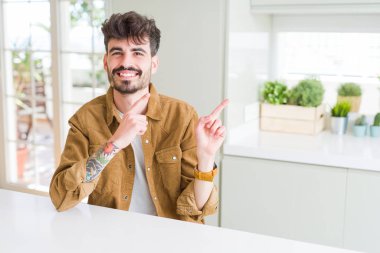 Sıradan bir ceket giyen genç adam beyaz masada oturuyor gülümsüyor ve kameraya bakıyor. İki eliyle ve parmaklarıyla kenarı işaret ediyor..