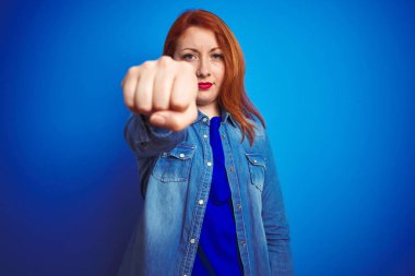 Genç, güzel, kızıl saçlı, kot gömlek giyen, mavi arka planda duran, yumruğunu yumruklayan, saldırgan ve öfkeli bir kadın, tehdit ve şiddet dolu.