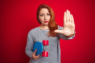 Kızıl saçlı genç bir kız, kırmızı izole edilmiş arka planda paten tutuyor. Açık elle dur işareti yapıyor. Ciddi ve kendinden emin bir ifadeyle, savunma hareketi yapıyor.