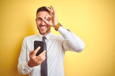 Genç yakışıklı iş adamı, sarı izole edilmiş arka plan üzerinde akıllı telefon kullanıyor. Gülümsüyor. Tamam işareti yapıyor.