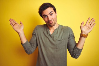 İzole edilmiş sarı arka plan üzerinde duran sıradan tişört giyen genç yakışıklı bir adam. Kolları ve elleri havada, kafası karışmış bir ifade. Şüphe kavramı.