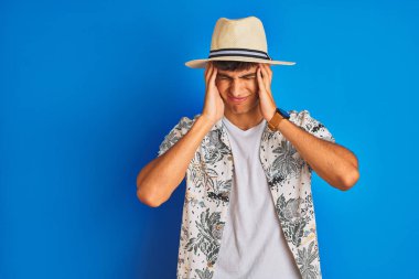 Tatilde olan Hintli bir adam, Hawaii gömlekli yaz şapkası takıyor, izole edilmiş mavi bir arka planı var. Baş ağrısı, acı ve migren yüzünden stres altında. Ellerini başının üstüne koy..