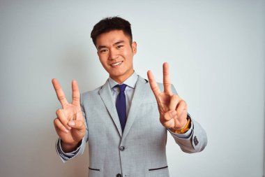 Asyalı Çinli işadamı takım elbise ve kravat takıp izole edilmiş sarı arka planda dikilip parmaklarını zafer işareti yaparken gösteren kameraya gülümsüyor. İki numara..