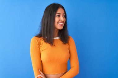 Turuncu tişört giyen genç, güzel Çinli bir kadın izole edilmiş mavi arka planda dikilip yüzünde gülümseme, doğal bir ifade ile yan yana bakıyor. Kendinden emin gülen.